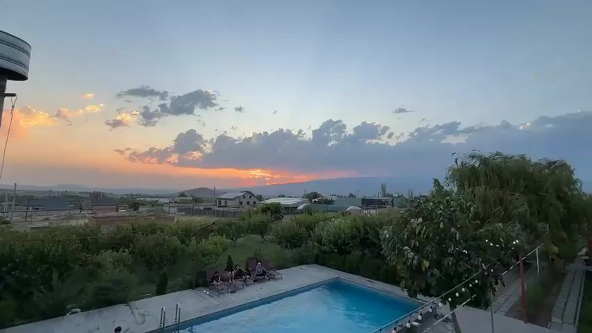 This video displays a panoramic view of a rural landscape at sunset. In the foreground, a rectangular swimming pool with clear blue water is visible. Several lounge chairs are placed around the pool, with four individuals seated on them. A large metal cylindrical object, possibly a water tank, stands to the left of the frame. Beyond the pool and lounge area, lush green trees and vegetation dominate the mid-ground. Scattered houses and buildings are visible in the distance, nestled within the trees. The background features rolling hills and mountains, with a vibrant sunset painting the sky with hues of orange, pink, and yellow. Clouds are scattered across the sky, with rays of sunlight piercing through them. The overall scene depicts a serene and picturesque setting at dusk.