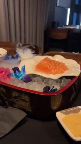 The video shows a close-up shot of a serving of salmon sashimi presented in a decorative bowl filled with ice. The salmon is a vibrant orange color and is sliced thinly, arranged on a white, flower-shaped plate. The bowl itself is dark brown with intricate gold detailing, possibly of pine trees. Blue and pink orchid-like flowers are placed around the salmon and on the ice. Wisps of smoke are visible, adding to the presentation. In the foreground, a small white triangular dish contains a yellow dipping sauce. The background is softly blurred, showing dark curtains and a wooden table or furniture, suggesting an indoor dining setting. The camera appears to be slightly tilted, focusing on the food presentation. There are no people visible in the frame.