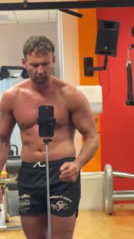 A shirtless, fair-skinned man with short, brown hair stands in a gym, wearing black shorts. He is holding a smartphone on a selfie stick, taking a video of himself. The man is flexing his arms and chest, and his facial expression is determined. The gym environment is visible in the background, with various exercise equipment and a red wall. The lighting is bright, and the overall impression is that of a fitness enthusiast documenting his workout.