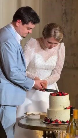 A man in a light blue suit and a woman in a white wedding dress are cutting a two-tiered white cake. The cake is decorated with berries and white dots. The couple is holding a knife together as they cut into the cake. They are both looking down at the cake. The environment appears to be indoors, possibly a wedding reception, with a wall in the background.