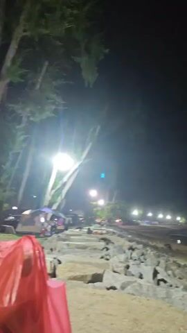 The video shows a nighttime scene at a beach. In the foreground, a large red plastic bag is prominent. Behind it, rocks and sand are visible. Further back, there are several tall trees. The middle ground reveals a more populated area with tents, some people, and motorcycles. In the background, there is a shoreline with lights lining the beach and reflections in the water. The overall atmosphere is dimly lit by artificial lights.