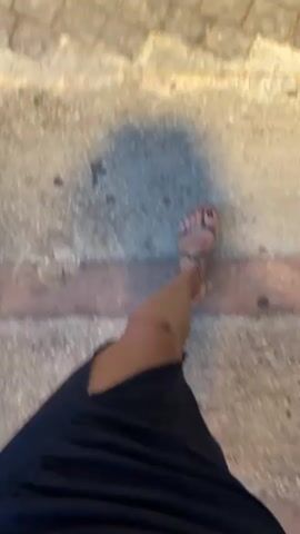 A first-person perspective video shows someone walking on a paved walkway. The person is wearing a dark dress and sandals. The walkway is bordered by a strip of red brick and then a patch of concrete. Beyond the concrete, there is a paved area with stone pavers, and further on, there are plants and trees illuminated by yellow lights. The scene appears to be outdoors, possibly in a garden or park. The video is shaky, indicating movement.