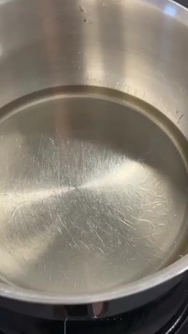 A close-up, high-angle shot shows a stainless steel pot filled with clear cooking oil. Five rectangular pieces of candy, resembling slices of bacon with red and white stripes, are dropped into the hot oil. As they begin to cook, the candy pieces curl and bubble in the oil.