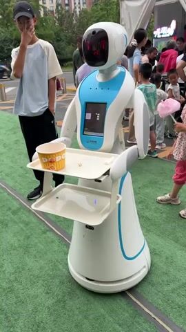 A white and blue robot with a screen on its chest stands on a green surface. It has two trays attached to its arms, one holding a yellow cup. A young girl in a floral dress and pink shorts is reaching out to the robot's tray. A teenager in a light blue t-shirt and black pants stands to the left of the robot, looking towards the camera. In the background, a crowd of people are gathered, some holding food items like cotton candy. The scene appears to be outdoors, possibly at an event or market, with buildings and a tent visible.