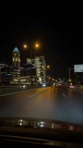 The video shows a nighttime view from inside a vehicle driving on a highway. The road is wet and reflects the headlights of the car and streetlights. On the left side of the road, there are several modern, illuminated skyscrapers. The buildings are tall and feature a variety of architectural styles, with different colored lights, including blue and yellow, accentuating their features. A car passes by in the opposite direction, its taillights visible. The sky is dark and clear, indicating it is nighttime. The camera perspective suggests the video is shot from the driver's or front passenger's seat.