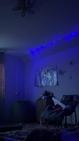 The video shows a room with blue string lights draped along the ceiling. A window is on the left side of the room, with a sheer curtain. Outside the window, it appears to be daytime. There are plants on the windowsill. In the center of the room, there is a dark-colored beanbag chair. To the right of the beanbag, there is a wooden cabinet. In front of the cabinet, a person is seated in a chair, partially visible. The person is wearing dark pants and a red and white striped shirt. They are holding a phone or remote in their right hand. The overall lighting of the room is dim and atmospheric, primarily from the blue string lights and the light coming from the window.