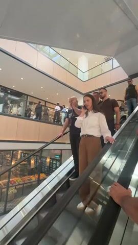 In a modern, multi-level shopping mall in Bjeliši, Montenegro, a woman with long dark hair, wearing a white shirt and brown pants, is seen descending an escalator while engrossed in a phone conversation. She is accompanied by several men, one in a brown polo shirt and sunglasses standing behind her, and another in a black t-shirt also on a phone call. An older man in a striped polo shirt is also on the escalator in the foreground.

The mall is brightly lit and bustling with numerous shoppers on various levels. The lower level features several retail spaces, including one with "organic" visible in its signage. On the ground floor, green and white balloons are part of a display, and a silver car or car display is visible among the crowd. The scene captures the dynamic environment of a busy shopping center.