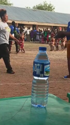 The video begins with a focus on a clear plastic water bottle on a green plastic table. Behind the bottle, two women stand, one in a gray t-shirt and black pants, and the other in a blue shirt that reads "Planning Committee" with a yellow skirt. A crowd of people is visible in the background, including children wearing traditional patterned clothing, seemingly participating in a dance. A rope on the ground marks the area where the dancers perform. The camera then shifts, focusing on the scratched surface of the green plastic table. The video is shot outdoors in what appears to be a rural setting.