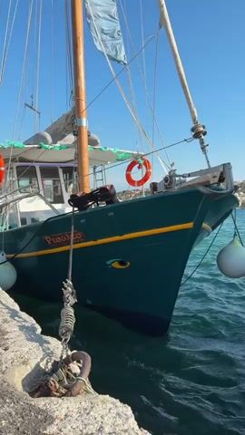A close-up shot of a green boat docked beside a stone jetty. The boat has a wooden mast with a partially furled sail and is secured by ropes and fenders. A life preserver is visible on the side of the boat. The water is choppy, and the sky is clear and blue. The name "Proua" is visible on the side of the boat.