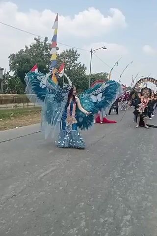 A woman in a detailed blue and teal costume with large, feathered wings adorned with butterflies is walking down a paved road. She is also wearing a long, shimmering blue dress that flares out at the bottom and has a silver crown on her head. The woman is smiling and looking to her right as she walks. In the background, there are other people in costumes, some of whom are carrying flags or poles. Trees and a blue sky with clouds are visible behind the road. The scene appears to be a parade or festival.