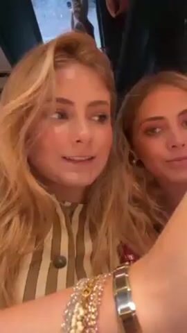 A woman in a striped top holds up an oyster for the camera, then eats it. She is indoors and has blonde hair. There is a second woman in the background.