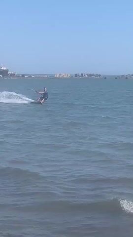 A kitesurfer is on the water. The kitesurfer is wearing a dark shirt and shorts, and a life vest. The kitesurfer is on a board and holding a kite string. The water is choppy, and there are waves. In the background, there are buildings and mountains. The sky is blue and clear. Another kitesurfer is in the distance, and there are several kites in the air. The video is shot from a low angle, close to the water.