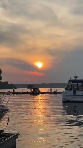 The video shows a sunset over a body of water. The sun is visible in the sky, and its rays are reflecting on the water. There are several boats docked on the water, and a line of trees can be seen in the distance. The sky is filled with clouds, and the overall scene is peaceful and serene. The video is shot from a low angle, looking out over the water.