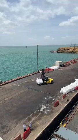 A yellow forklift is shown on a dock, lifting two large white bags. The forklift is positioned near the edge of the dock, with the sea visible in the background. There are several other white bags stacked on the dock to the right. Two people in light-colored clothing are standing on the dock to the left, near the forklift. The sky is cloudy. The sea is a teal color. The dock is made of asphalt.