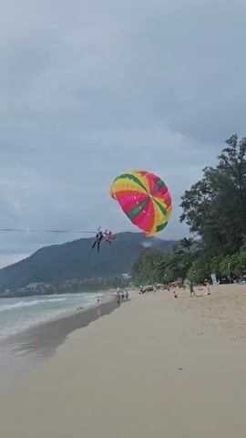 Two people are parasailing above a sandy beach with a calm ocean. The parasail is colorful, with segments of red, pink, yellow, green, and blue. The parasailers are harnessed together and appear to be enjoying the ride. In the background, there is a line of trees and some hills, suggesting a tropical or coastal location. The sky is overcast with some patches of blue visible. There are a few people walking along the beach, and beach chairs are set up in the sand. The overall scene depicts a leisure activity at the beach.