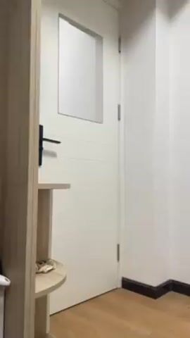 Here is a detailed description of the video:

The video shows the corner of a room. We can see a section of a light brown parquet floor with a dark trim along the wall. The walls are painted white. There's a white door with a small, opaque rectangular window near the top. The door has a black handle. To the left of the door, there's a light wood cabinet or shelf. The camera pans to the right, revealing a dumbbell and parts of a pillow on the floor along the wall.