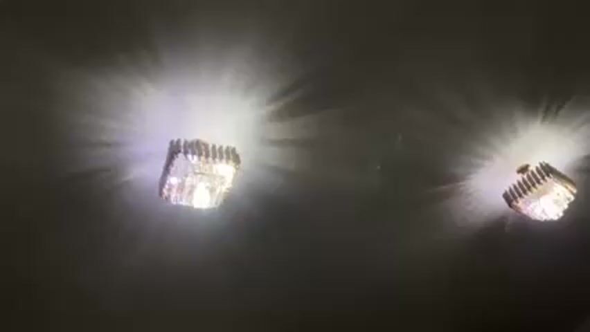 The video shows two illuminated light fixtures attached to a ceiling. Each fixture is square and has a gold frame, with rows of clear, square, glass-like pieces. The light from the fixtures radiates outward in a starburst pattern.