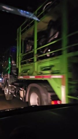 The video shows a nighttime view from inside a vehicle. The camera is positioned to capture a perspective that is rotated. In the foreground, there is a green metal structure, possibly part of a vehicle or equipment. Beyond this, the road is visible, illuminated by streetlights and the headlights of other vehicles. Traffic appears to be present, with the red taillights of cars visible in the distance. The environment is dark, with the artificial lights creating streaks and reflections. There is no clear indication of people or specific actions taking place.
