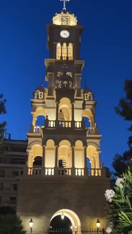 A clock tower illuminated at dusk. The tower is tall and ornate, with multiple levels of arches and decorative elements. A clock face is visible at the top. The sky is a deep blue, indicating twilight. In the foreground, there is foliage and a fence. The tower is lit with warm yellow lights, highlighting its architecture. The overall scene suggests a peaceful and historic setting.