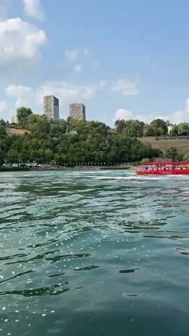 A low-angle, vertical shot captures a bright, sunny day with a clear blue sky dotted with white clouds. The foreground is dominated by choppy, greenish-blue water, with small white bubbles and ripples on the surface. In the mid-ground, a long, narrow red boat with a white roof and many passengers is moving from left to right across the frame, leaving a white wake. Behind the boat, a scenic landscape unfolds. Lush green trees cover a hillside that rises towards the background. Several multi-story buildings, appearing to be apartments or hotels, are visible atop the hill, with some showing light-colored facades. Further in the background, on the right side of the frame, a section of a waterfall is visible, with white water cascading over rocks. The overall scene suggests a popular tourist destination, likely a river with a waterfall, where boat tours are offered.