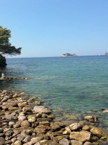 A person is snorkeling in clear, shallow water near a rocky shore. The person is wearing a white bikini and fins, and is looking down into the water. The sun is shining, and the sky is clear blue. In the distance, there are a few small islands and some boats.