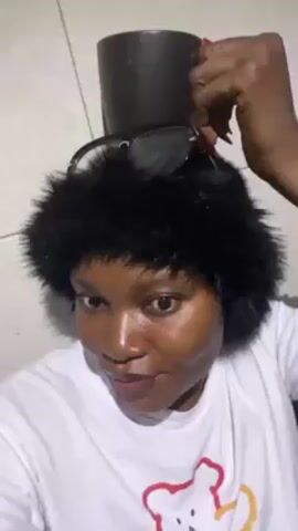 Here is a description of the video:
A woman wearing a white T-shirt with a cartoon bear design and a black faux fur hat with sunglasses on top is holding a black mug on her head. She is looking at the camera. The background shows tiled walls.