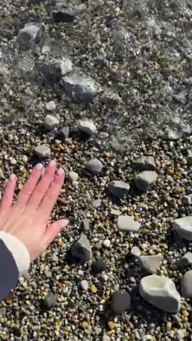 The video shows a hand interacting with water on a rocky beach. The hand has pale skin and manicured nails. It reaches out and touches the water as it washes over the pebbles and larger rocks on the beach. The water is clear and shallow, and the rocks are various shades of grey and black. A small portion of a blue and white jacket sleeve is visible in the lower left corner of the frame. The beach appears to be sunlit.