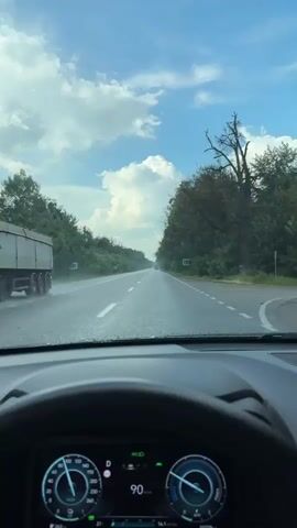 The video shows a first-person view from inside a car driving down a highway. The steering wheel and a digital dashboard are visible in the foreground. The dashboard displays the car's speed, which fluctuates between 90 and 88 km/h, and other vehicle information. The road ahead is a two-lane highway lined with trees and vegetation on both sides. The sky is partly cloudy with a bright blue sky visible. There are road signs visible along the side of the road. The overall scene depicts a drive through a rural or suburban area on a clear day.