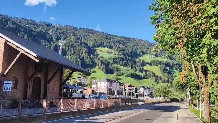 This is a video of a small European village nestled in a valley. The village is surrounded by green, tree-covered mountains under a clear blue sky. In the foreground, there is a brick building on the left with a dark roof and an overhang. Next to it, there is a fence and some parked bicycles. A road runs through the middle of the frame, leading towards the village. The village itself consists of several buildings, some of which appear to be houses and shops. There are some cars parked along the road. A cable car line is visible ascending one of the mountains. The trees on the right side of the frame are lush and green. The overall scene suggests a peaceful and scenic rural setting.