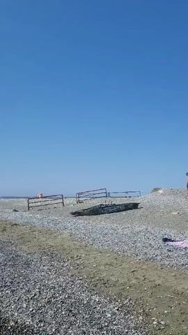A person is seen walking on a beach near a yellow car. The beach is covered in pebbles and sand. In the background, there is a clear blue sky. There are some metal structures on the beach, possibly remnants of a fence or other beach equipment. The person is wearing swim trunks and appears to be alone on the beach. There is also a towel and a bag lying on the sand. The person walks towards a piece of debris on the beach and appears to be examining it. The video is shot from a distance and is slightly shaky.