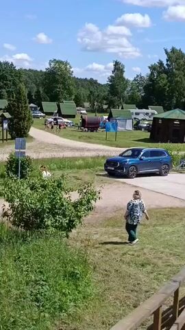 A blue SUV is parked at a campsite. A person walks away from the SUV. In the background, there are small cabins and trees. The sky is blue with scattered clouds.
