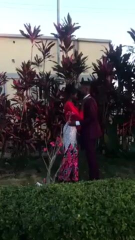 A couple is embracing in front of a shrub.  The woman wears a red blouse with a long, patterned skirt in colors including white, red, and black. The man wears a maroon suit with a white shirt.  They are both Black.  They stand in front of tall leafy plants with reddish leaves, in front of a low building. The sky is light blue.