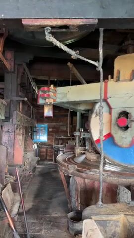This is a low-angle interior shot of a large, old, wooden machine, possibly a mill. A large, worn, wooden wheel with blue and red painted segments is visible on the right side of the frame, partially obscured by a thick rope. The wheel is mounted on a wooden axle supported by wooden beams. Various gears, ropes, and wooden structures are visible throughout the machine, suggesting a complex mechanical system. The overall environment appears rustic and industrial, with wooden beams and walls. There are no people visible in the video.