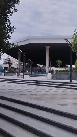This video shows a wide shot of an outdoor public space with modern architecture and landscaping. The camera pans from left to right, revealing an elevated walkway or bridge structure supported by white cylindrical pillars. Below the walkway, a dark, open space is visible, with more pillars and some greenery. In the foreground, there are wide, grey steps leading up to a paved area with a geometric pattern of hexagonal and square tiles. Several black, modern lampposts are placed strategically throughout the scene. To the right, a grand entrance archway with white columns and a decorative, green-patterned ceiling is prominent, featuring the words "DEHOON BOZORI" in large, dark lettering. Beyond the archway, trees and what appear to be fountains can be seen. A few people are scattered throughout the scene, some sitting or walking near the entrance and on the elevated walkway. The sky is overcast, suggesting a cloudy day.