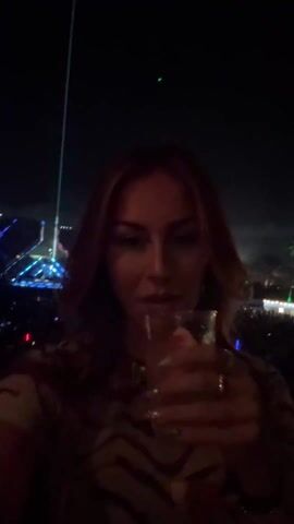 Here is a description of the video:
A woman is enjoying an outdoor concert at night. She is holding a small glass of clear liquid, possibly a drink, and she takes a sip while facing the camera. Behind her is a large crowd of people, and beyond them is a stage with bright lights and a large screen displaying the names "Afrojack" and "Niki." The stage is surrounded by beams of light, enhancing the concert atmosphere.