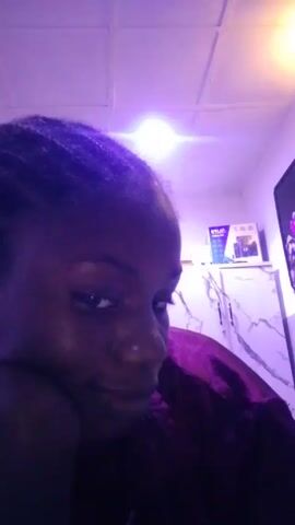 A dark-skinned woman with braided hair is close to the camera, looking at it from an angle. She smiles briefly. She is wearing a shiny purple shirt. The room is lit with a purple glow. Behind her is a white cabinet with a package on top, and a framed image on the wall. The ceiling is white and tiled. Loud music is playing.