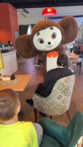A full-body costume character, appearing to be Cheburashka, stands in a dimly lit indoor venue. The character has large, furry brown ears and a beige face with large, round eyes and a small orange bow tie. It is facing the camera, with its arms outstretched. In the foreground, two people are seated, facing away from the camera, looking towards the character. The person on the left is wearing a yellow t-shirt. The person on the right is wearing a black shirt and is seated in a chair with a patterned upholstery. The background shows several other people, including children, and what appears to be a buffet table with decorations. The floor is made of polished wooden planks, and the ceiling has red circular lights. The overall impression is that of a children's party or event.