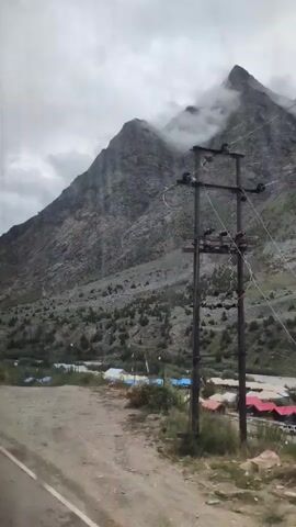 A cloudy, overcast day shows a mountainous landscape. The foreground features a dirt road and a grassy verge. A yellow sign with black lettering is visible on the left. In the midground, a cluster of blue and white tents is situated in a valley. The background is dominated by steep, rocky mountains, with some areas covered in mist. The camera is in motion, suggesting it is mounted on a vehicle traveling along the road.