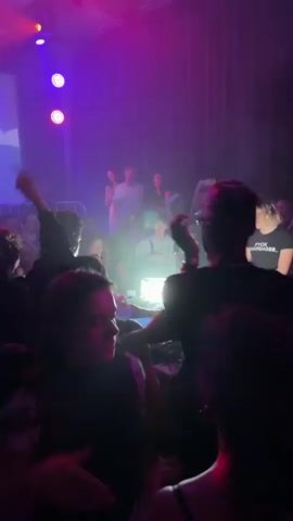 Here’s a description of the video:

A crowded indoor scene, likely a nightclub or concert venue, is captured under bright, colorful lighting. The foreground shows the backs of several people, suggesting the camera is positioned within the crowd. A DJ booth is visible, with a person in a cap operating the equipment. Behind the booth, there's a raised platform or seating area filled with more people. Some are standing and raising their arms, seemingly enjoying the music and atmosphere. The lighting is predominantly pink and purple, with beams shining down, and there appears to be some fog or haze in the air.