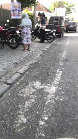 A still frame shows a street scene with several motorcycles and a small truck. In the foreground, a portion of a motorcycle handlebar and frame is visible. To the left, a red motorcycle is parked on a brick-patterned sidewalk. Further back, a woman wearing a headscarf and a floral dress stands near the red motorcycle, facing away from the camera and appearing to interact with it. To the right of the woman and slightly further back, a black motorcycle is parked, and a person wearing a helmet and a patterned shirt is seated on it, facing the camera. Behind the black motorcycle, a flatbed truck is parked. The environment appears to be outdoors, with a paved road in the foreground and a sidewalk. Trees and buildings are visible in the background. The lighting suggests it is daytime, possibly late afternoon or early evening.