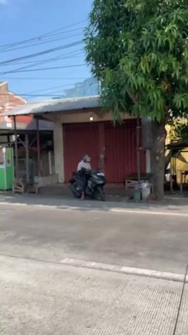 A person wearing a white cap and light-colored shirt is sitting on a black motorcycle parked on a paved road. The person appears to be looking towards a closed shop with a red corrugated metal shutter. A large tree with green leaves stands to the right of the shop. The environment appears to be an outdoor urban or semi-urban setting with buildings and power lines visible in the background. The overall lighting suggests it is daytime.