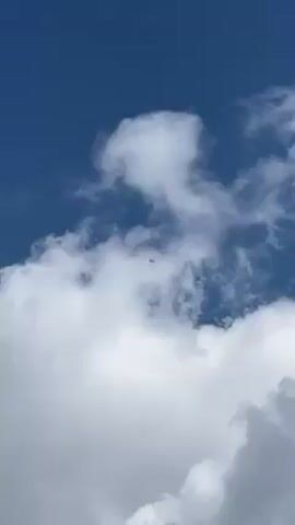 The video shows a low-angle view of a clear blue sky with fluffy white clouds, and a distant aircraft moving from the left to the right of the frame. The plane is a small dark shape against the blue background.