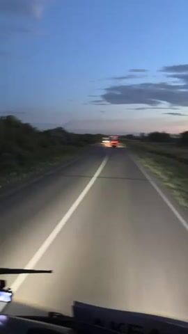 This is a first-person perspective video taken from the inside of a vehicle, likely a truck or bus, driving on a paved road at dusk or dawn. The vehicle's headlights illuminate the road ahead, casting bright white beams. Two trucks are visible in the distance, their headlights and taillights glowing. The road is marked with a white line down the center. The surroundings consist of sparse vegetation and trees on either side of the road. The sky is a deep blue with some wispy clouds. The camera is mounted on the dashboard, showing the windshield, a portion of the dashboard, and the wiper blades. The video is short and shows the vehicle continuing its journey along the highway.