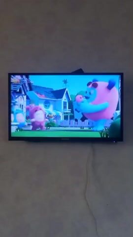 The video shows a television screen displaying a scene from an animated children's show. On the screen, there are five characters. From left to right, there are three small, round, blob-like characters: one green, one blue, and one purple. To their right stands a larger, pink, blob-like character with blue accents on its horns and ears. This pink character is holding a smaller, pink, blob-like character in its left arm. All characters appear to be in an outdoor setting with a house and trees in the background. The scene seems to be from a moment of dialogue or action, as some characters have their mouths open. The television is mounted on a wall with a textured finish. A black cable is visible running down from the top of the television.