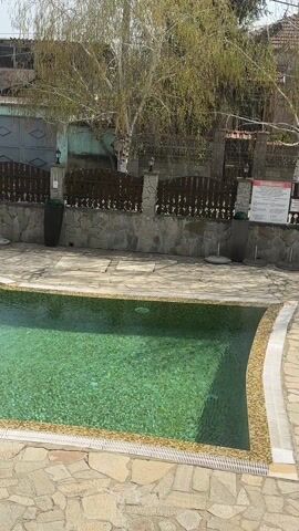 The video shows an outdoor swimming pool area on a sunny day. The pool is irregularly shaped with a smaller, round, jacuzzi-like section attached. The water is a green hue. The pool is surrounded by stone paving. A low stone wall with a decorative wooden fence runs along one side of the pool area. There are large dark planters placed along the wall. Trees and buildings can be seen in the background. A metal ladder leads into the pool. The camera pans slightly upward and to the left, revealing more of the surrounding scenery. No people are visible in the shot.