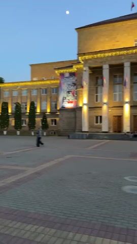 The video begins with a view of a large, lit building with columns and flags. A person walks across a paved courtyard in front of the building. The camera pans to the right, showing more of the building and a few trees. The scene then shifts to a wider view of the courtyard, with people visible in the distance and mountains in the background. The sky is a gradient of twilight colors.