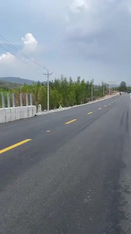 The video depicts a drive along a paved road with yellow lines. The landscape features green trees and hills in the distance under a cloudy sky. A vehicle passes on the road ahead, while power lines run parallel to the road. The camera is likely mounted on a moving vehicle, capturing the view from its perspective.