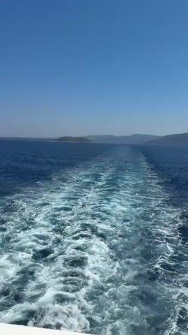The video shows a wake behind a boat in the ocean. The water is a deep blue, and the sky is clear and blue. In the distance, there are islands or a coastline. The wake is white and foamy, indicating the boat is moving at a good speed. The camera is positioned on the back of the boat, looking out over the water.