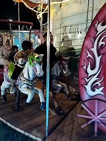 The video shows a carousel in motion, likely at a fair or amusement park at night. A young child rides a white horse, accompanied by an older male, presumably a father or guardian. Other people are riding on the carousel, including a woman in a hijab and a child in a crescent moon-shaped carriage. The carousel is decorated with lights, and the surrounding environment is visible in the background, showing tents, other people, and general fairground activity.