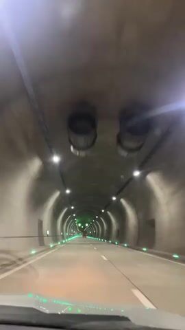 The video shows a first-person perspective of driving through a tunnel. The tunnel walls are concrete and curved, illuminated by overhead lights and green lights along the sides of the road. There are traffic lights with green arrows pointing downwards visible in the distance. The road ahead has white lane markings.